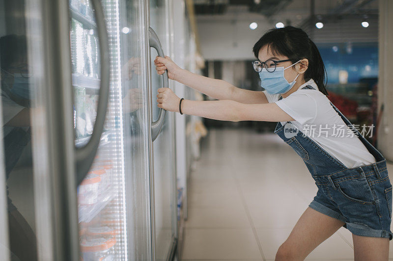 An asian chinese young girl looking at the refrigerated section choosing for her ice cream pulling opening the refrigerator door